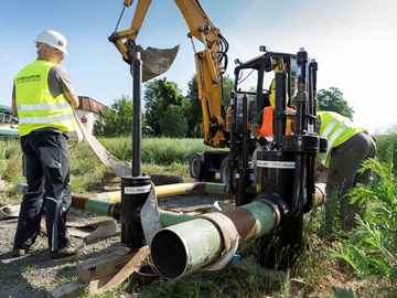Drei Mitarbeiter und ein Bagger bei Arbeiten an Rohrleitungen auf einer Wiese Drei Mitarbeiter und ein Bagger bei Arbeiten an Rohrleitungen auf einer Wiese